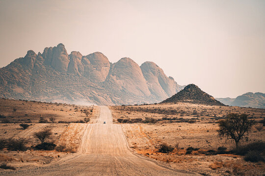 View of a long, straight dirt road disappearing into the horizon, leading towards jagged mountains under a pale sky, Namibia, Namibia.