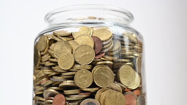 Close-up video of a transparent glass jar rotating on itself filled with euro coins of different denominations. Concept of savings, finance, economy and money accumulation with dynamic motion.