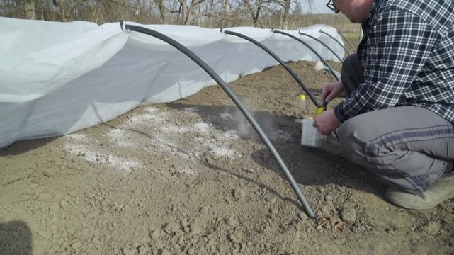 A man in a checkered shirt kneels near a row of soil covered with agrofibre and uses a shovel to apply ash, demonstrating the process of planting crops in a rural area
