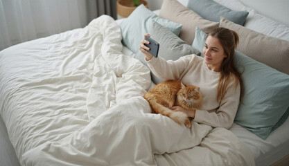Obraz na płótnie Canvas Woman sitting on bed and taking selfie with cat in cozy bedroom 