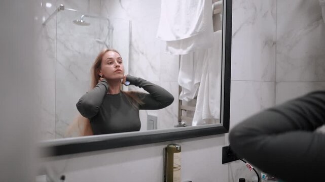 A beautiful and young girl in a bathrobe does morning self-care, dries her hair with a hairdryer after a shower, in the morning in front of a mirror in the bathroom