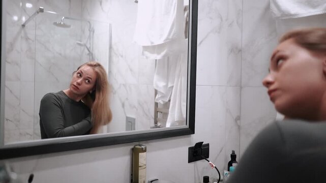 A beautiful and young girl in a bathrobe does morning self-care, dries her hair with a hairdryer after a shower, in the morning in front of a mirror in the bathroom