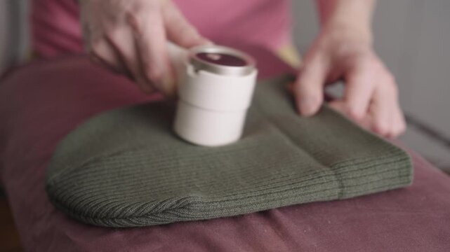 Man using lint remover appliance for fabric care, cleaning and restoring knitted winter hat from pilling and fuzz, demonstrating textile maintenance for clothing quality, close-up, slow motion.
