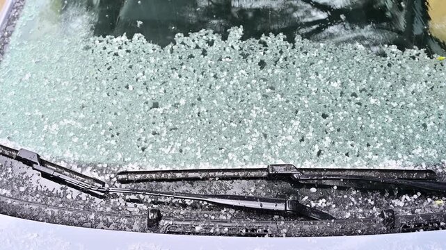 Close-up view from a parked car showing the windshield during an intense hailstorm. Hailstones hitting and accumulating on the glass create a dramatic weather scene with impact