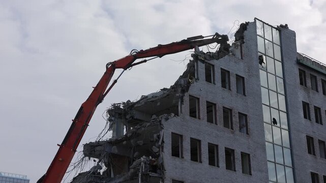 A giant demolition crane tears down an office building piece by piece, its powerful claw gripping and dismantling each section with force.