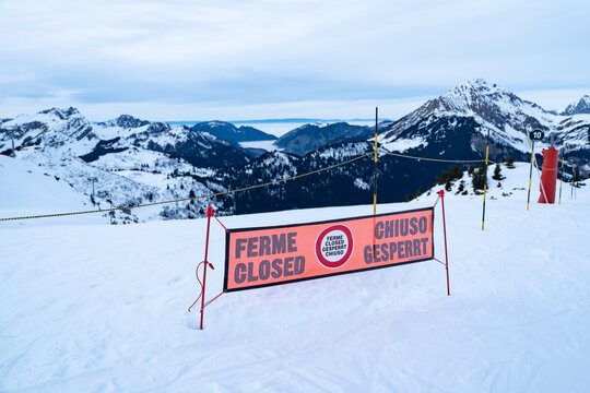 View of a closed ski run with a bright orange sign displaying "Ferme Closed Chiuso Gesperrt" against a backdrop of snow-covered mountains, Val-d'Illiez, Valais, Switzerland.