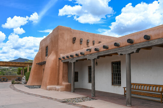 View of a traditional adobe building with exposed wooden beams under a bright blue sky dotted with fluffy clouds, Santa Fe, New Mexico, United States.