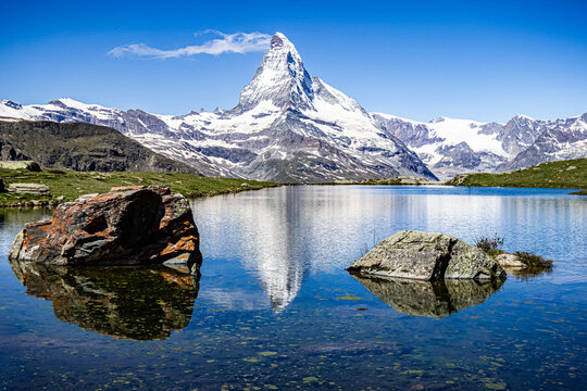 View of the majestic Matterhorn mountain reflected in the tranquil Stellisee lake, framed by rugged rocks and a clear blue sky, Zermatt, Valais, Switzerland.