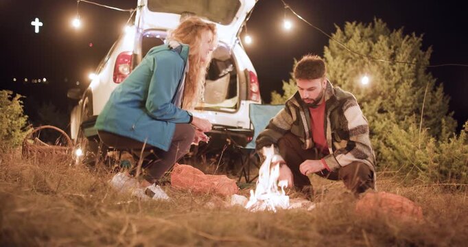 Couple enjoys campfire cooking at night in outdoor location with camper van around warm fire and scenery under bright lights in a natural setting