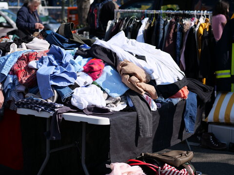 A high-angle, eye-level shot captures a white folding table overflowing with a messy pile of secondhand clothing at a sunny outdoor flea market. The assortment includes denim, cotton shirts, and color