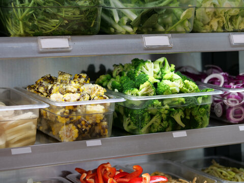 A high-angle, medium-close-up shot features a refrigerated salad bar or buffet display with fresh, chopped vegetables. Clear plastic containers are filled with vibrant green broccoli florets, yellow