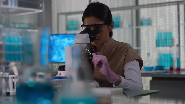 Female scientist in protective gear working in a modern laboratory to test human DNA samples. Looking into the microscope. Selective focus on the person