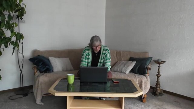 Senior woman multitasking using laptop and smartphone on sofa at home