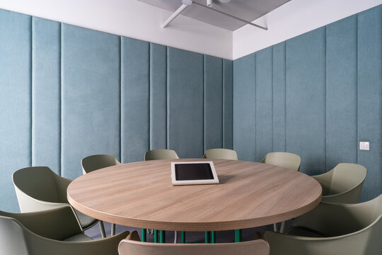 Modern meeting room with round wooden table, upholstered acoustic wall panels and minimalist chairs, contemporary office interior design with tablet on table