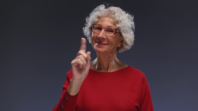 Old woman shows a forbidding gesture with wag finger. Senior female shakes her hand in glasses looking at camera, forbids, it is impossible, rejection. Strict order or Stop sign. Wagging or shake.