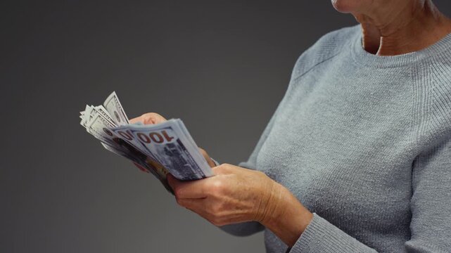 Unrecognizable old mature woman counts cash dollars, holding bundle one hundred dollar banknotes. Arms of senior female holds paper USD money in wrinkled hands after winning lottery bonus. Close up