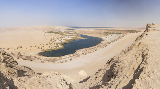 View of a serene, dark lake nestled amid the undulating golden dunes of a vast desert landscape under a clear sky, Faiyum Governorate, Egypt.