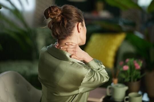 A woman seen from behind massages her neck to soothe muscle tension and stress while sitting in a peaceful, plant-filled living room.