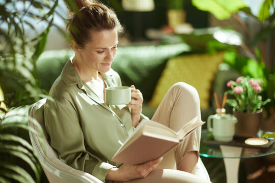 A woman enjoys a slow living moment, holding a warm mug and her favorite book while sitting in a sunlit, plant-filled room designed for rest and mental wellness.