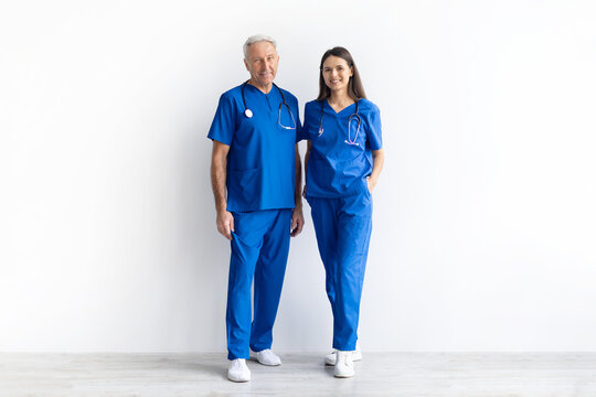 Full length portrait of two healthcare professionals in blue scrubs standing on a white studio background. Experienced senior male doctor and female medical worker showing teamwork in healthcare