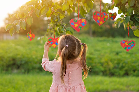 A girl decorates a tree by hanging hearts from pieces of multicolored puzzles, symbolizing awareness of autism, support and care for people with autism spectrum disorder.