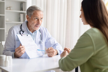 Obraz premium Senior male doctor showing medical document to female patient in clinic. Healthcare professional explaining treatment plan while patient points at information on clipboard