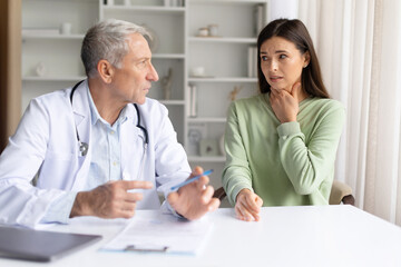 Obraz premium Female patient touching throat while explaining symptoms to doctor during medical consultation in clinic. Healthcare professional listening and assessing possible throat infection