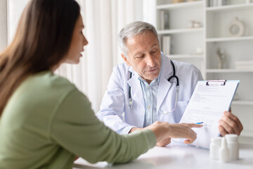Fototapeta premium Senior male doctor explaining medical document and treatment plan to female patient during clinic consultation. Healthcare professional reviewing healthcare paperwork