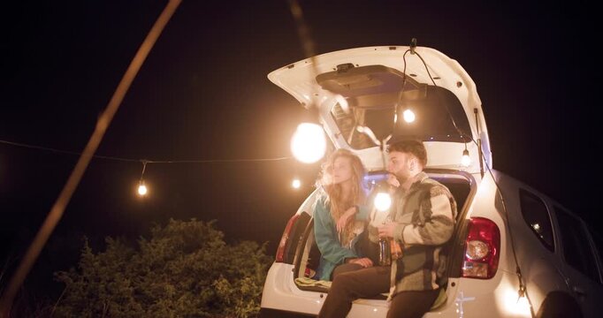 Couple enjoys night under string lights while relaxing at the back of their car in a quiet location during a clear evening