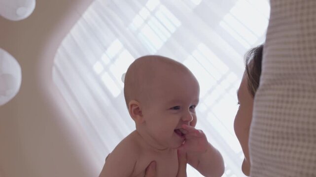mother lifting baby above chest on bed. morning exercise and play mom with infant with bright window light. cheerful movement and eye contact. happy domestic maternal routine. cozy bonding