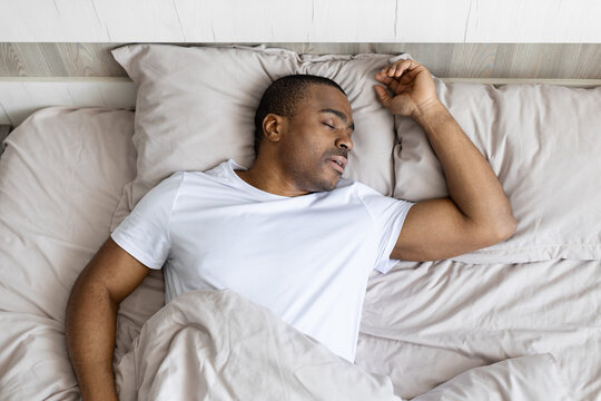 Young black man sleeping peacefully in bed during daytime, relaxed posture and calm expression, representing healthy rest, recovery and comfortable lifestyle in home bedroom setting