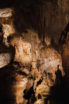 View into a deep limestone chamber of Petralona Cave in Halkidiki, Greece, with stalactites and stalagmites emerging from shadow and warm spotlighting.
