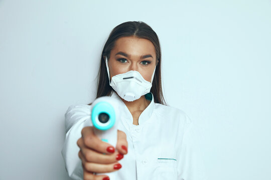 Healthcare worker points a contactless thermometer toward the camera while wearing a mask in a well-lit indoor setting during the COVID-19 pandemic