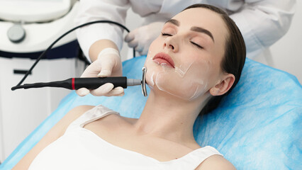 Caucasian female patient receiving RF lifting facial treatment in a cosmetology clinic, with a technician applying a handheld device on her face © wedmoments.stock