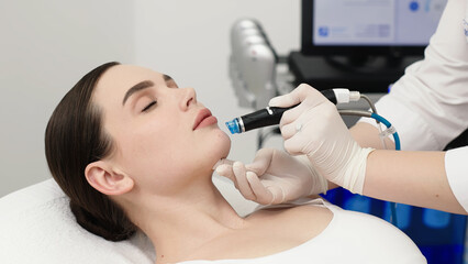Female patient receiving Hydrafacial treatment in a modern beauty clinic, technician using a handheld device for skin rejuvenation, clinical setting visible in background © wedmoments.stock