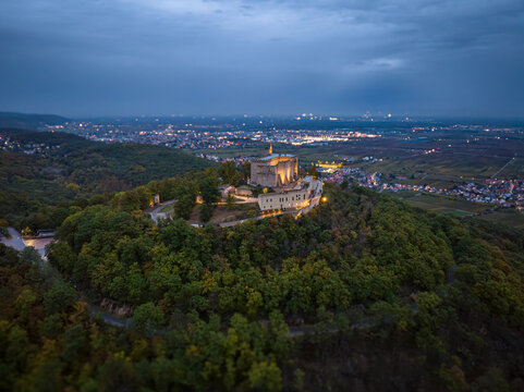 Aerial view of Hambach Castle atop a hill surrounded by lush forests, with city lights twinkling in the distance, Neustadt an der Weinstrasse, Rheinland-Pfalz, Germany.