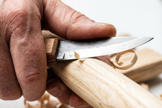 Hands using a sharp carving knife to whittle a light wooden stick producing thin curly shavings