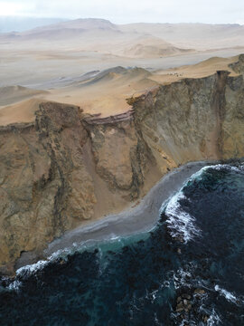 Aerial view of dramatic cliffs plunging into the dark turquoise sea with a small beach nestled at its base, contrasting with the arid desert landscape, Paracas, Ica, Peru.