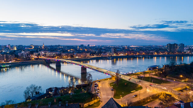 Aerial view of a radiant bridge reflecting the twilight sky over the still waters of the Danube, contrasting with the city lights of Petrovaradin, Vojvodina, Serbia.