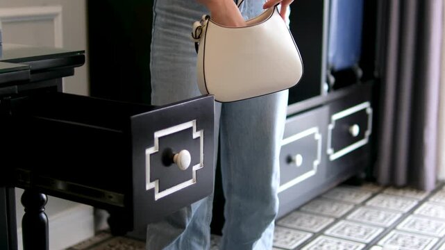 Girl in jeans takes a hairbrush and items from her handbag, placing cosmetics and personal belongings into black drawer with white pattern on stylish wooden table.