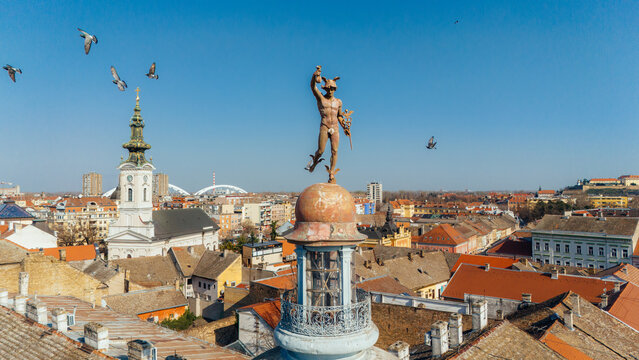 Aerial view of a statue atop a building, pigeons flying around, with the city stretching out in a tapestry of red roofs and sun-kissed walls, Novi Sad, Vojvodina, Serbia.
