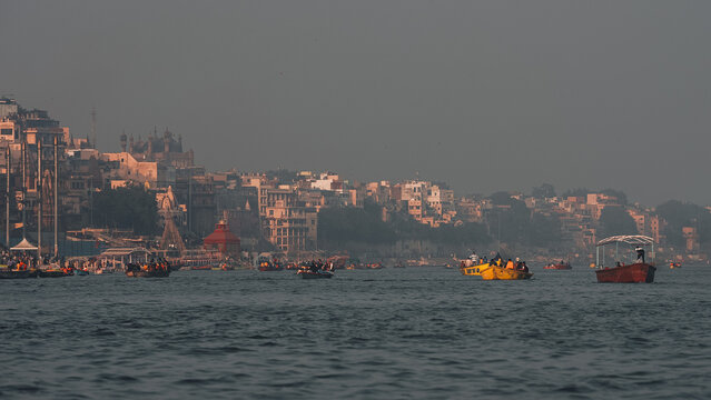 View of boats glide on the Ganges River against the backdrop of ancient temples and buildings under a hazy sky, Varanasi, Uttar Pradesh, India.