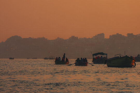 View of boats gliding across the shimmering water under a hazy, orange sky, silhouettes of buildings fading into the horizon, Varanasi, Uttar Pradesh, India.