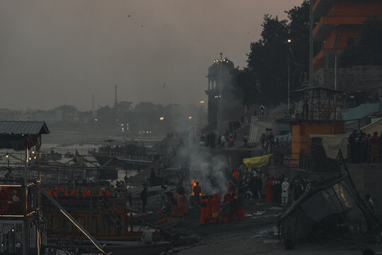 View of funeral pyres blazing, smoke rising amidst crowds near the riverbank, with boats lining the shore, under a dusky sky, Varanasi, Uttar Pradesh, India.