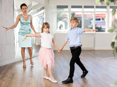 Two smiling kids, boy and girl learning cha-cha under watchful eye of female dance instructor in bright choreography studio..