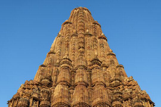 View of an ancient temple reaching towards the clear blue sky, crafted with intricate carvings and warm earthy tones, standing as a testament to history, Khajuraho, Madhya Pradesh, India.