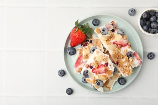 Pieces of tasty yogurt bark with berries, almond flakes and granola on white tiled table, flat lay. Space for text