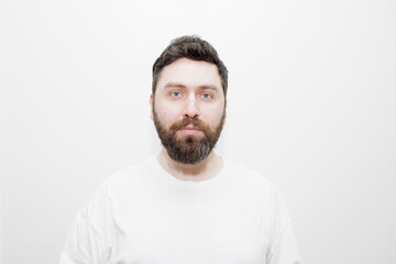 Obraz premium A direct, neutral headshot portrait of a man with dark hair, beard and blue eyes in a white t-shirt on a white background. Stock photo