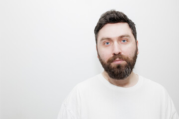 Fototapeta premium A direct, neutral headshot portrait of a man with dark hair, beard and blue eyes in a white t-shirt on a white background. Stock photo