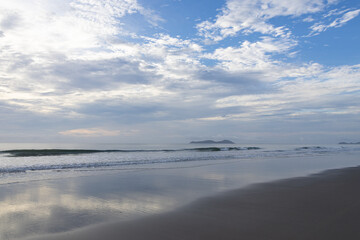 Fototapeta premium Serene coastal landscape with gentle waves on wet, reflective sand under a soft, cloudy sky. 